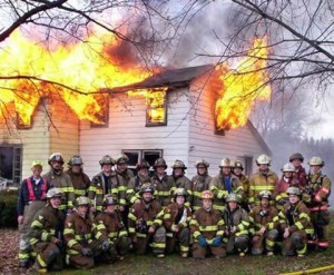 A large group of firemen are posing for a group photo in front of a house that is on fire.